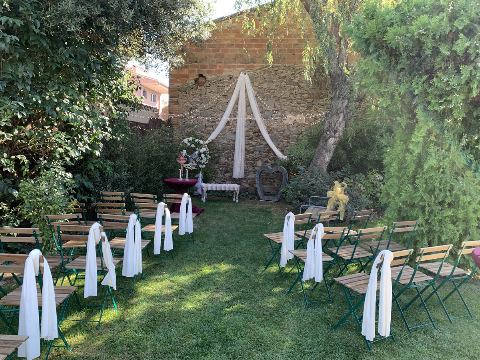 Cadires amb xals blancs sota els arbres del jardí, amb l'arc de teles a la paret de pedra