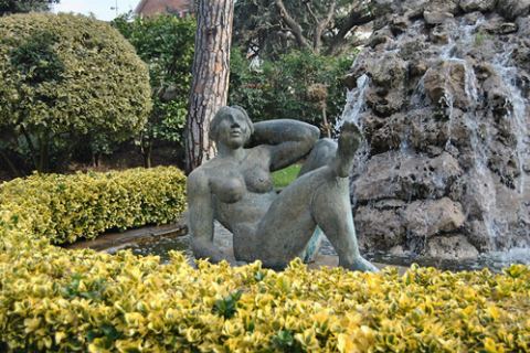 Escultura d'una dona recolzada davant la cascada, entre arbustos grocs